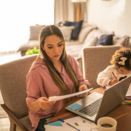 Jeune femme assise devant un ordinateur et lisant un papier. Sa fille assise à côté d'elle en train de dessiner. Illustre une mère en train de faire le dossier MDPH de son enfant